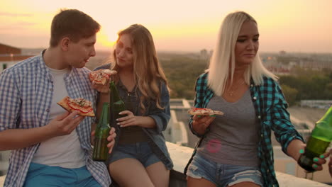Two-couples-in-love-eat-pizza-and-drink-beer-on-the-roof.-Girls-shyly-look-at-their-boyfriends.